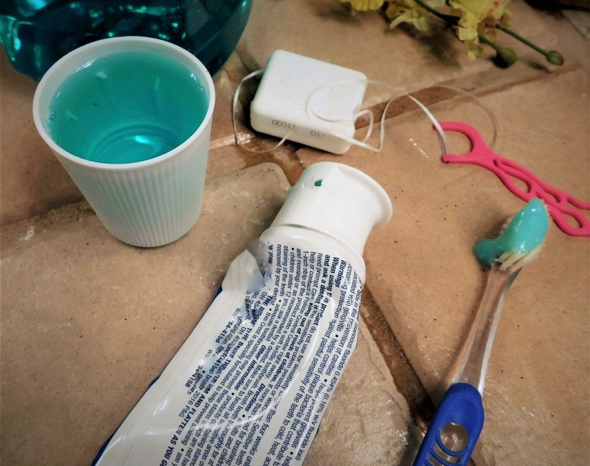 A cup of mouthwash, tube of tooth paste and container of floss sitting on a bathroom